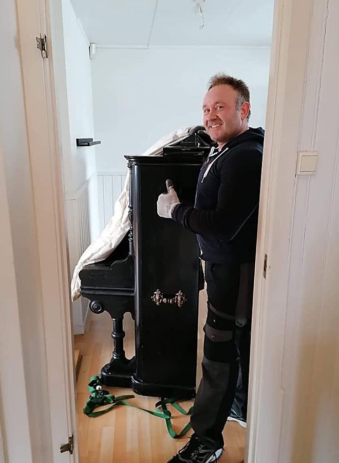 Ansatt bærer et piano ut gjennom en dør, sikret med stropper.